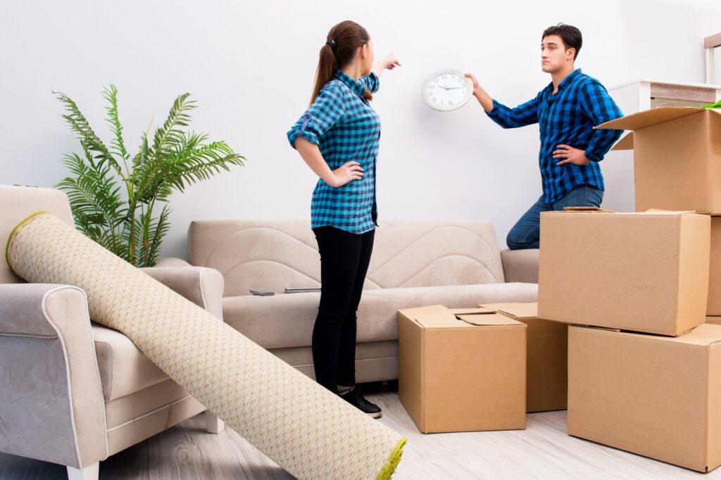 A couple is organising their new house after their DIY move and is surrounded by cartons and rolled carpet